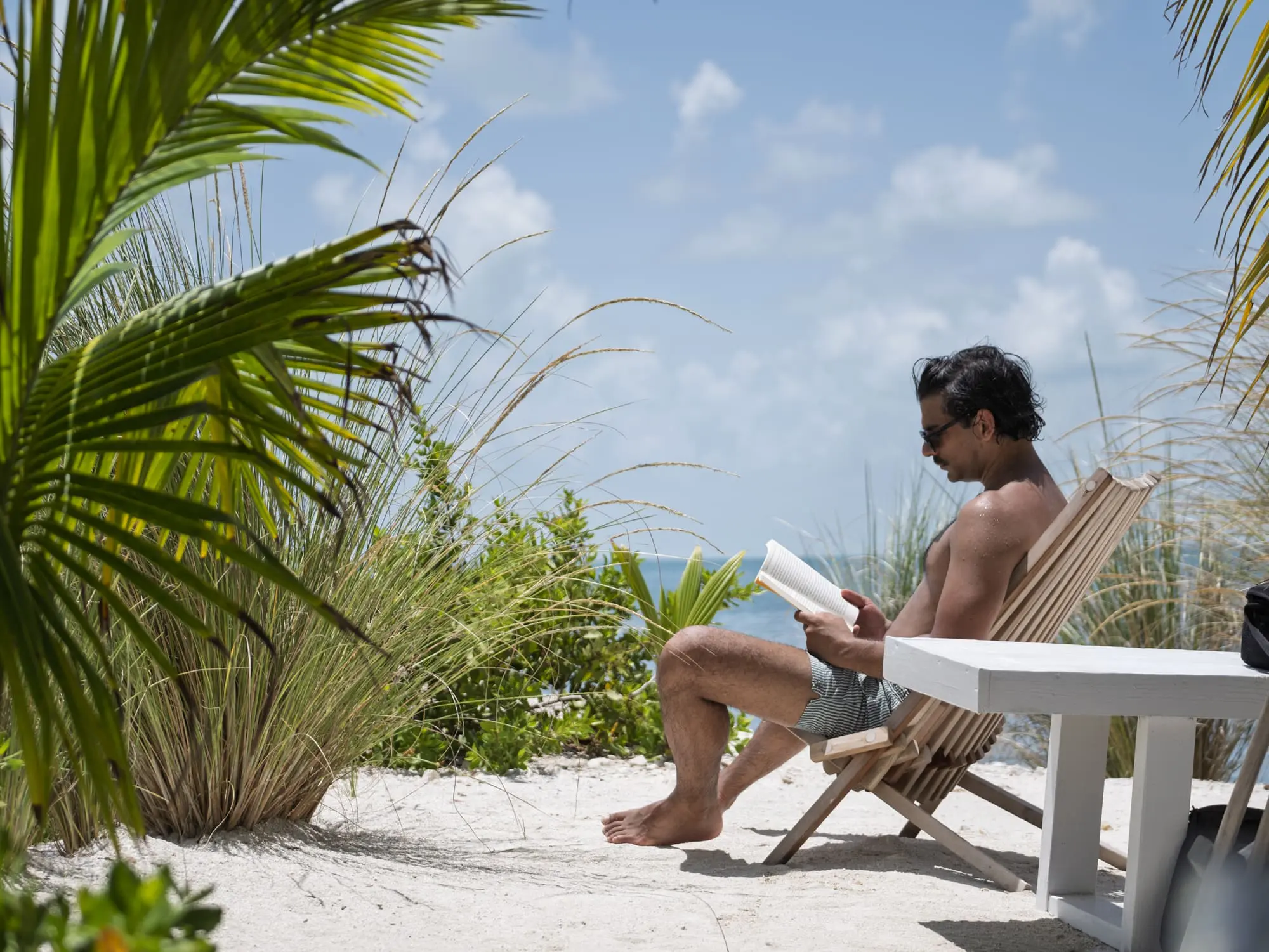 Image of a man reading a book in an adirondack style chair next to the ocean